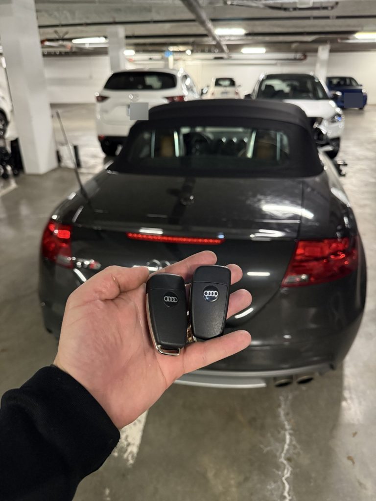 Hand holding two Audi key fobs behind a black Audi convertible in a Vancouver parkade