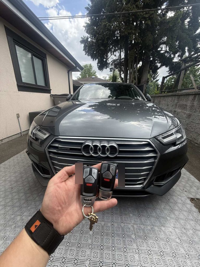 Hand holding two Audi key fobs in front of a grey Audi in a Vancouver driveway