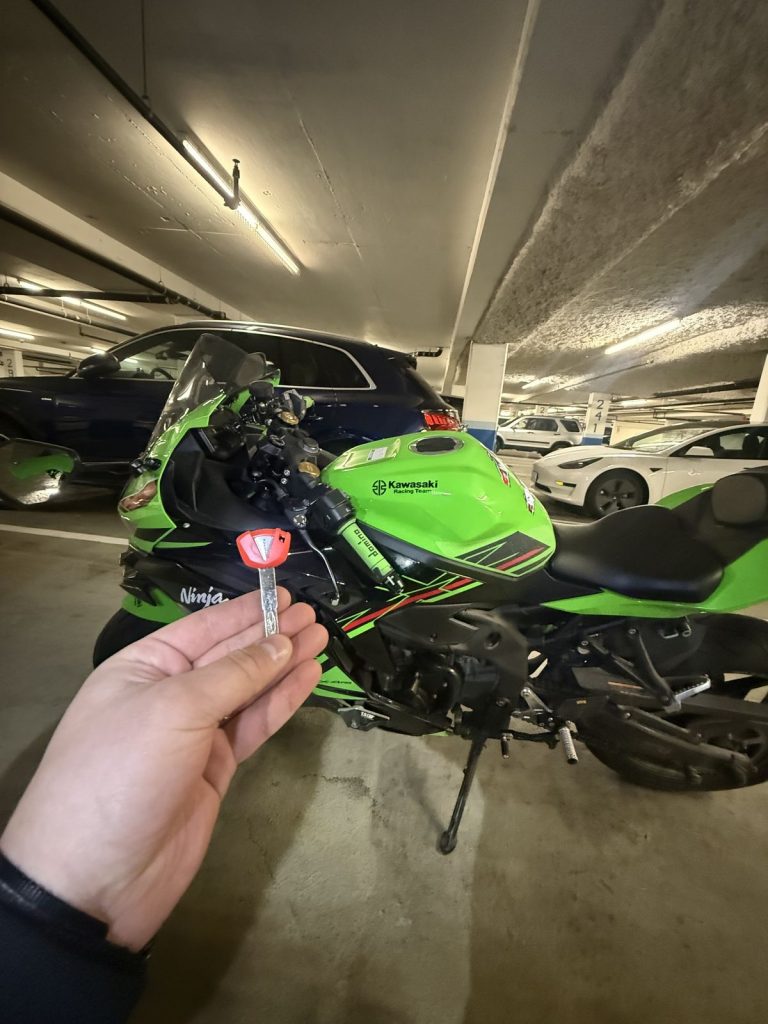 Hand holding a red motorcycle key in front of a green Kawasaki Ninja in a Vancouver parkade