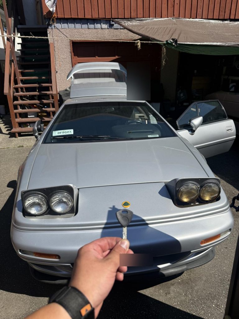 Hand holding a Lotus key in front of a silver Lotus Esprit in Vancouver