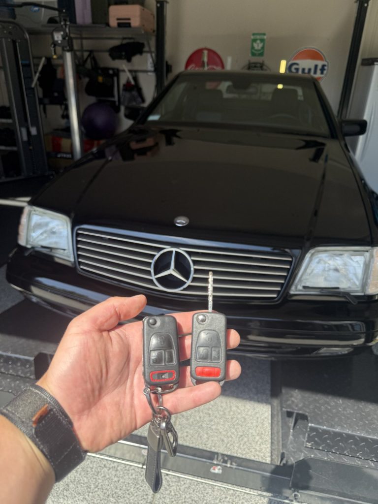 Hand holding two Mercedes-Benz flip keys in front of a classic black Mercedes-Benz SL in a Vancouver garage