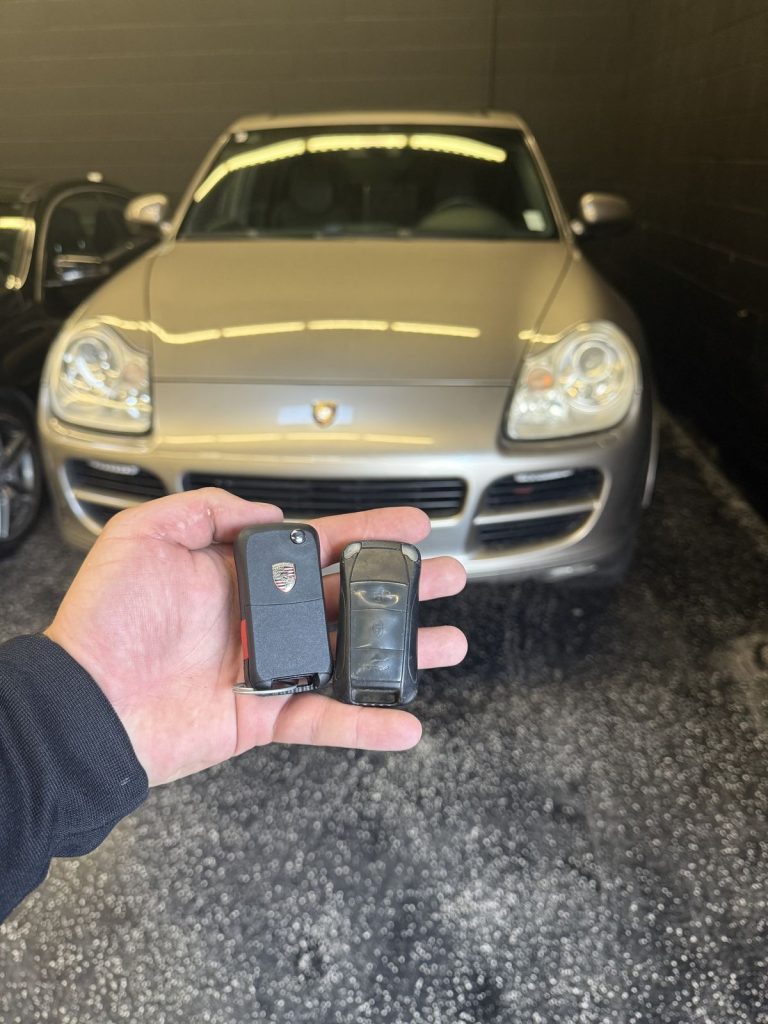 Hand holding two Porsche key fobs in front of a silver Porsche Cayenne in a Vancouver garage